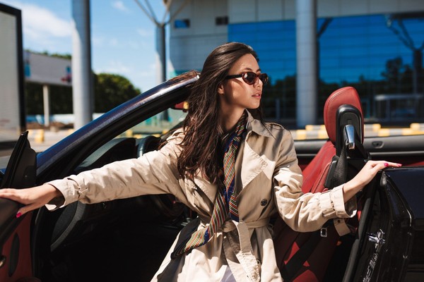 young-pretty-lady-sunglasses-trench-coat-opening-cabriolet-car-door-while-dreamily-looking-aside-with-airport-background